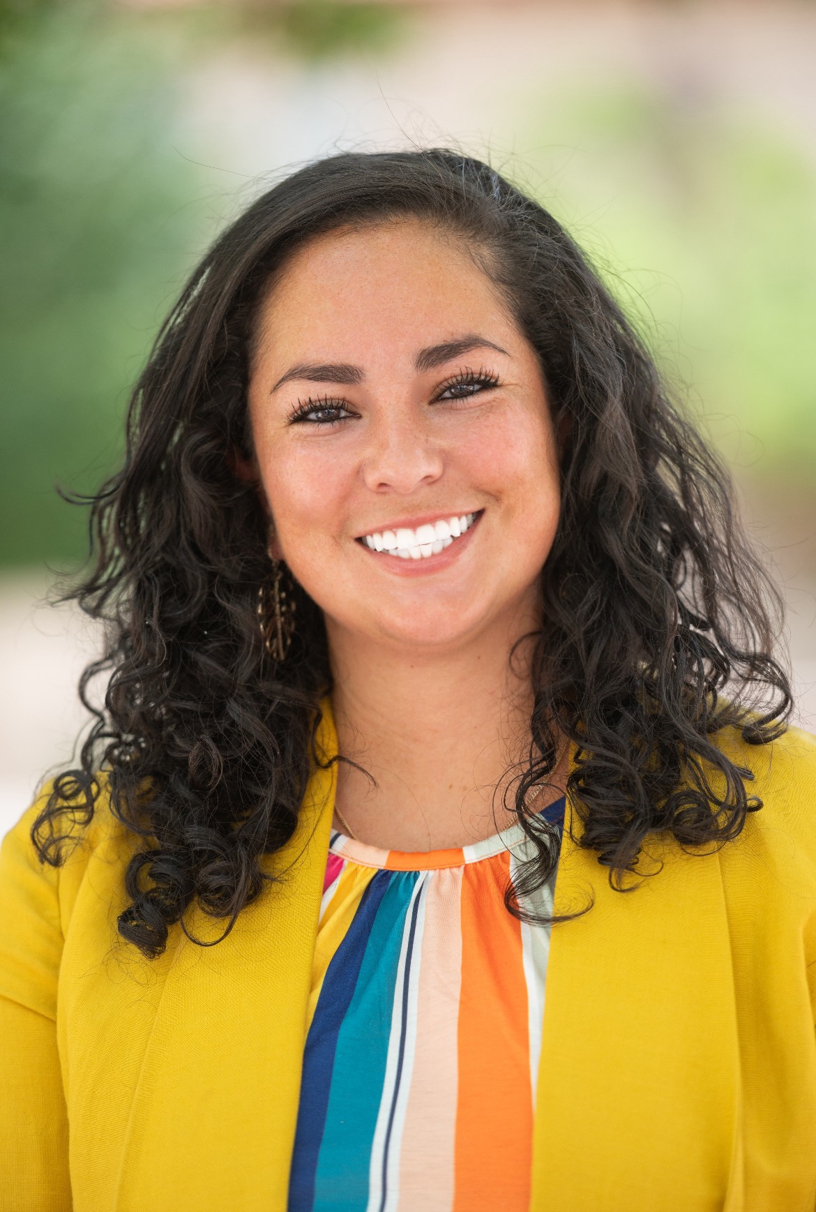 The image is a portrait of a woman smiling directly at the camera. She has long, curly dark hair that falls over her shoulders. She is dressed in a vibrant yellow blazer and underneath it, she is wearing a colorful striped top featuring vertical stripes in shades of blue, orange, pink, and white. The background of the image is blurred with tones of green and light brown, likely suggesting an outdoor setting. The woman's expression is joyful, and her smile is broad, showing her teeth.